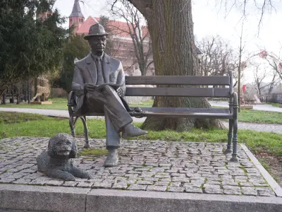 Brązowa rzeźba mężczyzny w kapeluszu na ławce z psem u stóp, w parku na Wzgórzu Tumskim w Płocku