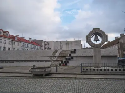 Pomnik 13 Straconych w Płocku z betonową ścianą pamięci, dzwonem w kręgu i tablicami, na tle zabudowy mieszkalnej zimą.