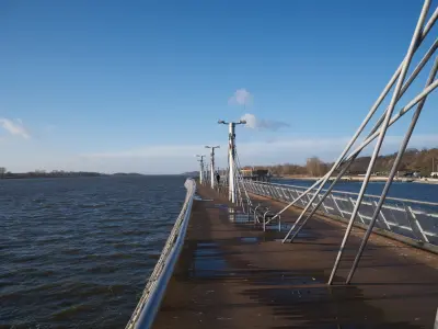 Drewniane molo nad Wisłą w Płocku z metalowymi masztami i ławkami, prowadzące w stronę rzeki przy słonecznej pogodzie