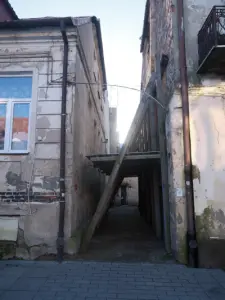 Wąskie przejście między zniszczonymi kamienicami w Płocku, z drewnianą konstrukcją podpartą belkami nad chodnikiem