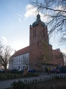 Ceglana wieża dzwonnicza bazyliki katedralnej w Płocku z zieloną kopułą, obok biały budynek i zaparkowane samochody zimą