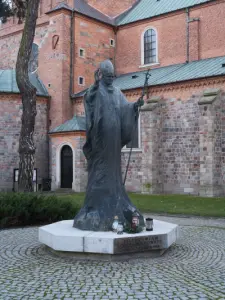 Pomnik Jana Pawła II na kamiennym cokole przed ceglaną katedrą w Płocku, z widocznym pastorałem i otoczeniem z brukowane
