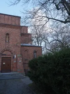Ceglany budynek Muzeum Diecezjalnego w Płocku z drewnianymi drzwiami, rzeźbą świętego i otaczającymi drzewami