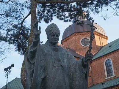 Pomnik Jana Pawła II przed ceglaną katedrą z kopułą i zielonym dachem w Płocku, otoczony drzewami